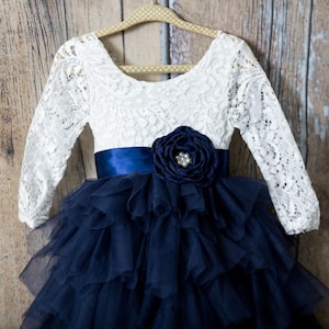 May include: A long-sleeved dress featuring a white lace bodice with a scoop neckline and a navy blue satin waistband. The waist is accented with a large navy blue fabric flower containing a central cluster of clear beads. The skirt consists of multiple layers of navy blue tulle ruffles.