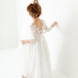 May include: A white lace dress with three-quarter length sleeves, a scalloped neckline, and a deep back cutout. The dress features a full-length skirt with a floral lace pattern and a solid white ribbon tied into a bow at the waistline.