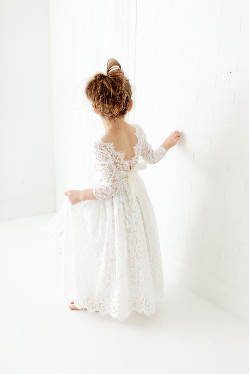May include: A white lace dress with three-quarter length sleeves, a scalloped neckline, and a deep back cutout. The dress features a full-length skirt with a floral lace pattern and a solid white ribbon tied into a bow at the waistline.