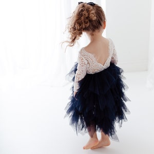May include: A dress featuring a white long-sleeved lace bodice with a deep V-shaped back and a voluminous, layered navy blue tulle skirt. The skirt consists of multiple tiers of ruffled fabric, creating a textured, full silhouette that falls below the knee.