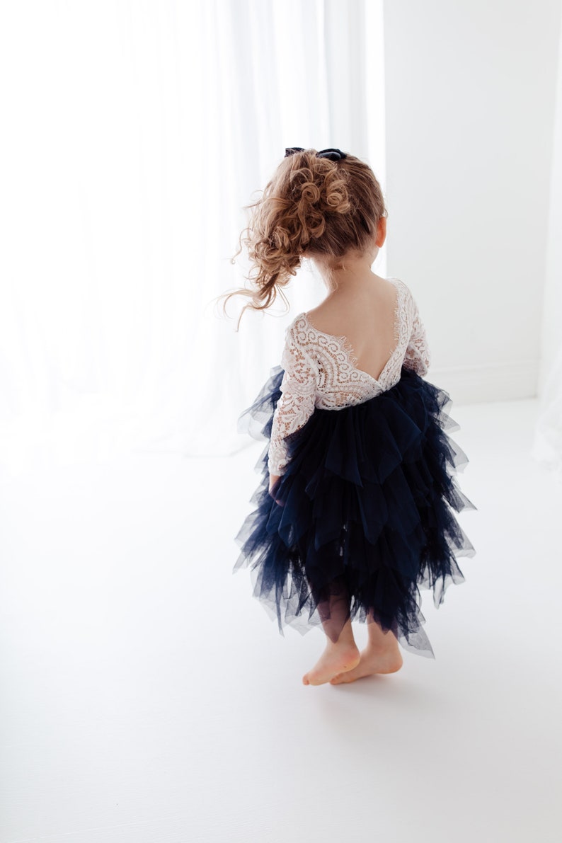 May include: A dress featuring a white long-sleeved lace bodice with a deep V-shaped back and a voluminous, layered navy blue tulle skirt. The skirt consists of multiple tiers of ruffled fabric, creating a textured, full silhouette that falls below the knee.