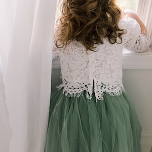 May include: A young girl wearing a white lace top and a sage green tulle skirt. She is standing by a window and looking out.