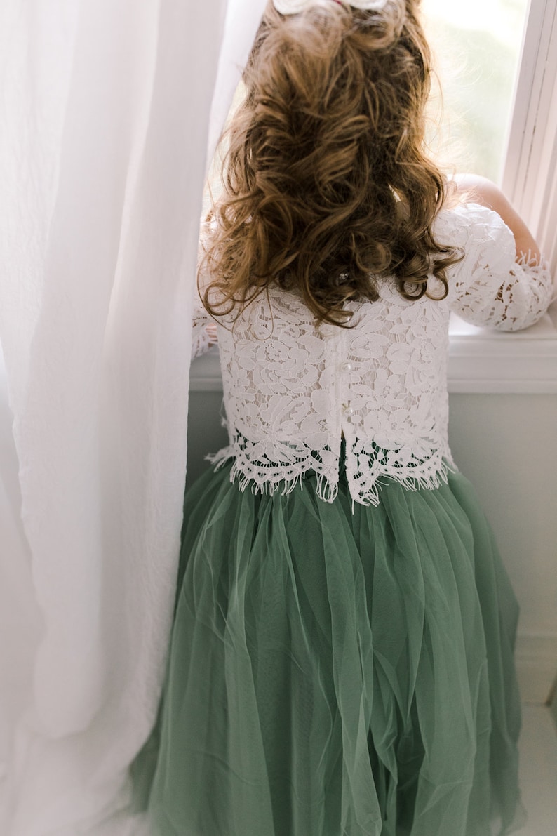 May include: A young girl wearing a white lace top and a sage green tulle skirt. She is standing by a window and looking out.