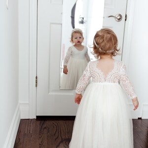 May include: A floor-length white dress with long lace sleeves and a sheer lace bodice featuring a scalloped back neckline. The skirt is made of layered tulle fabric. A matching white headband is worn with the dress, which is visible in a mirror.