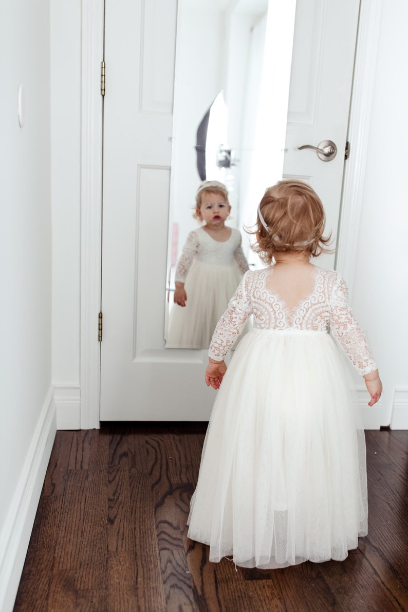 May include: A floor-length white dress with long lace sleeves and a sheer lace bodice featuring a scalloped back neckline. The skirt is made of layered tulle fabric. A matching white headband is worn with the dress, which is visible in a mirror.