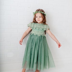 May include: A young girl wearing a sage green tulle dress with a lace bodice and a green leaf crown. The dress has a flowing skirt and the girl is standing on a white brick background.