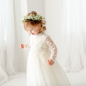May include: A long-sleeved white dress featuring intricate lace detailing on the bodice and sleeves, a high neckline, and a flowing skirt. The garment is paired with a floral crown made of small white blossoms and green leaves.