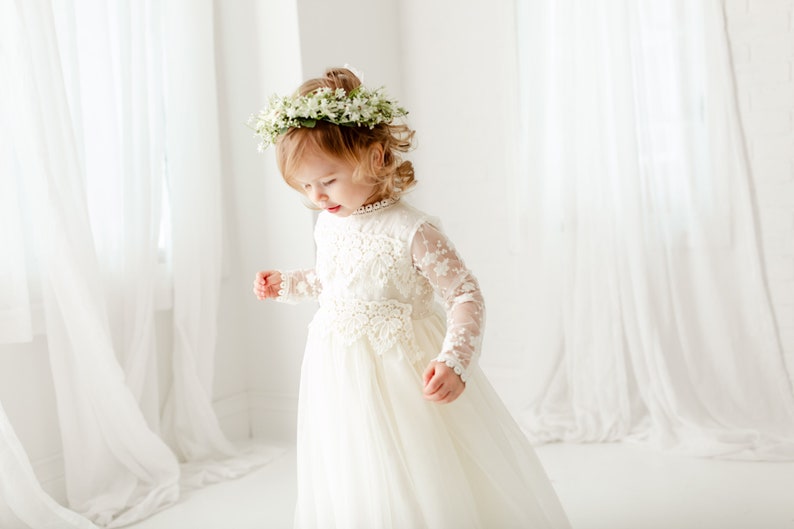 May include: A long-sleeved white dress featuring intricate lace detailing on the bodice and sleeves, a high neckline, and a flowing skirt. The garment is paired with a floral crown made of small white blossoms and green leaves.