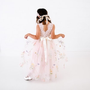 May include: A floor-length dress featuring a white lace bodice with a V-neckline and a sheer, pale pink tulle skirt adorned with colourful embroidered flowers and butterflies. A wide white satin ribbon is tied in a bow at the waist.