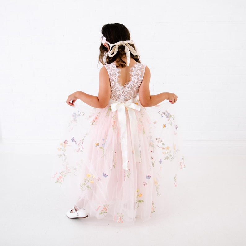 May include: A floor-length dress featuring a white lace bodice with a V-neckline and a sheer, pale pink tulle skirt adorned with colourful embroidered flowers and butterflies. A wide white satin ribbon is tied in a bow at the waist.