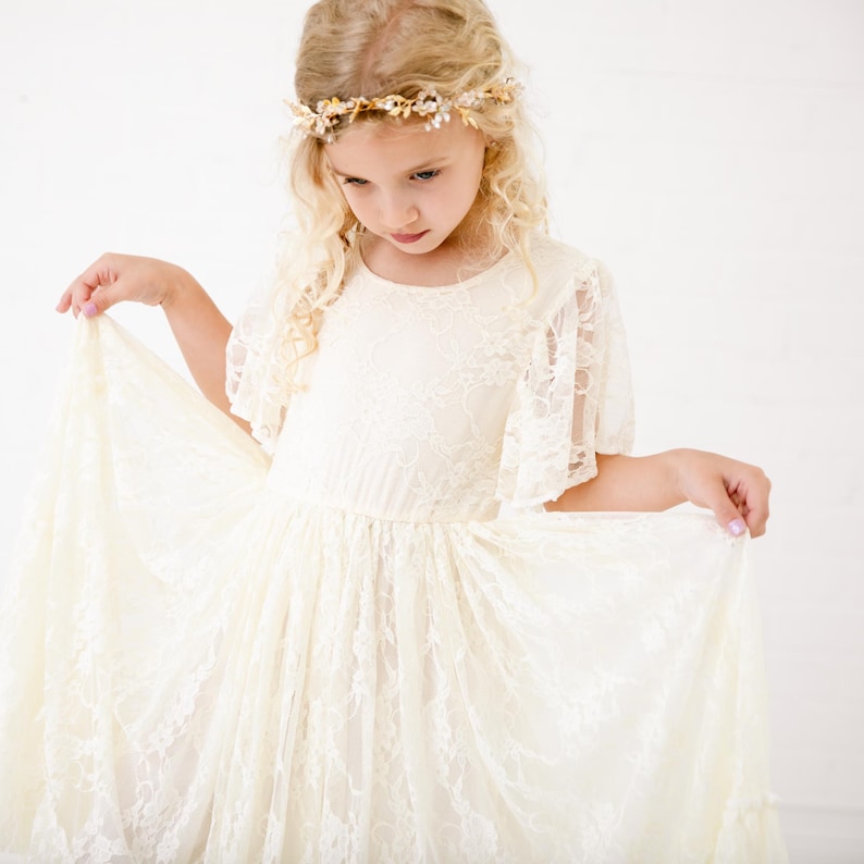 以下が含まれることがあります： 金色の花の冠をかぶった、白いレースのドレスを着た若い女の子。ドレスはフリルのあるネックラインと短い袖をしています。女の子はドレスのスカートを手に持っています。