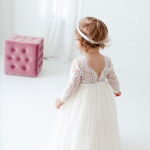 May include: A long-sleeved white dress featuring a lace bodice with a deep V-shaped back and a full, layered tulle skirt. The garment is paired with a simple white headband, creating a cohesive look for a formal occasion or special event.