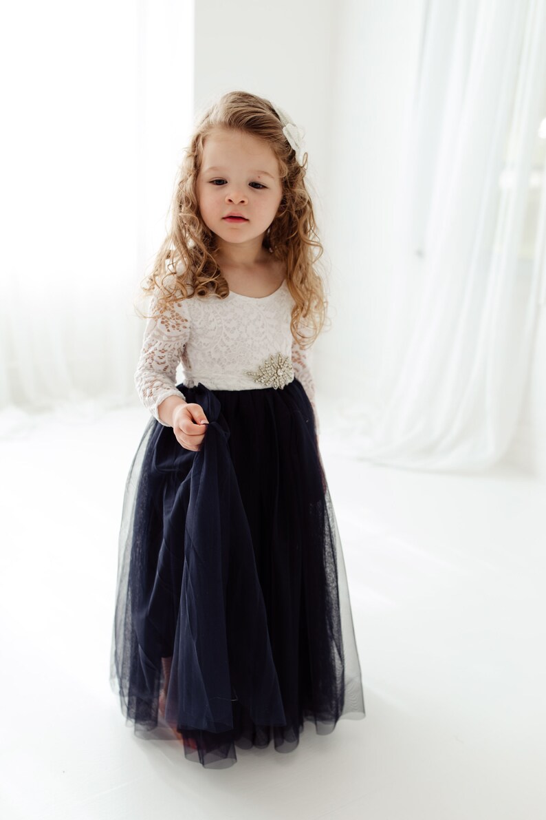 May include: A young girl wearing a white lace long-sleeved dress with a navy blue tulle skirt and a silver embellished belt.