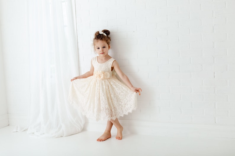 May include: A young girl wearing a white lace dress with a peach-colored flower accent at the waist. She is standing in front of a white brick wall.
