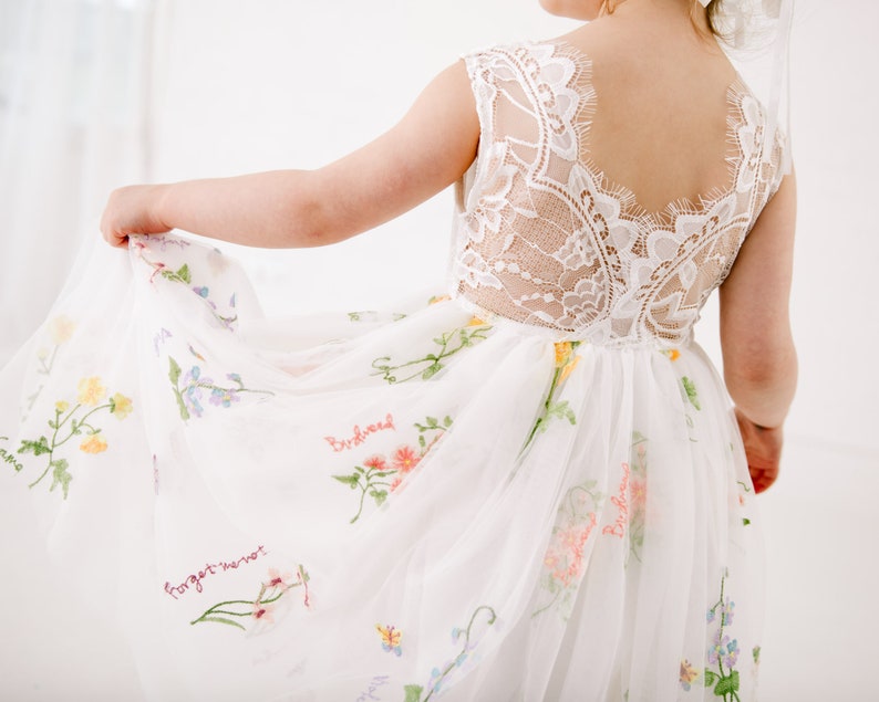 Puede incluir: Vestido blanco de tul con bordado floral y las palabras "no me olvides" y "semillas de p&aacute;jaro" bordadas en la falda. El vestido tiene una espalda de encaje.