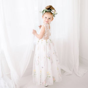 Puede incluir: Una ni&ntilde;a peque&ntilde;a con un vestido blanco de tul con flores bordadas y una corona de flores. El vestido tiene una espalda de encaje y la ni&ntilde;a est&aacute; de pie frente a una cortina blanca.