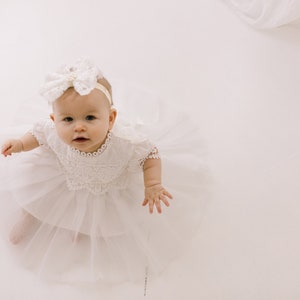 K&ouml;nnte beinhalten: Ein kleines M&auml;dchen in einem wei&szlig;en Spitzenkleid mit einem wei&szlig;en Schleifenhaarband. Das Baby schaut mit einem s&uuml;&szlig;en Ausdruck zur Kamera.