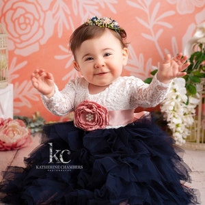 May include: A dress featuring a white long-sleeved lace bodice and a voluminous navy blue tulle skirt. A wide, dusty pink satin sash with a large matching fabric flower and pearl centre accents the waist. A floral headband adorns the head.