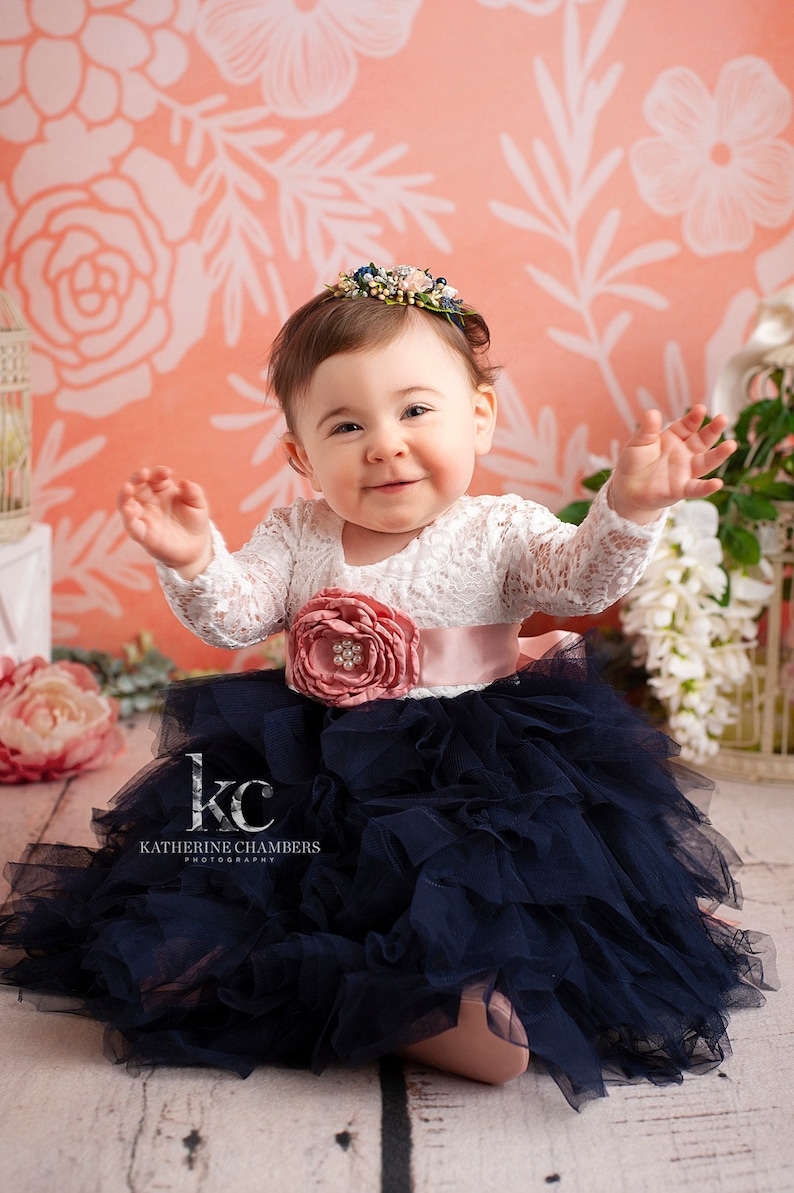 May include: A dress featuring a white long-sleeved lace bodice and a voluminous navy blue tulle skirt. A wide, dusty pink satin sash with a large matching fabric flower and pearl centre accents the waist. A floral headband adorns the head.