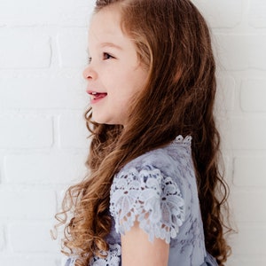 K&ouml;nnte beinhalten: Ein junges M&auml;dchen mit langen braunen Locken tr&auml;gt ein hellblaues T&uuml;llkleid mit Spitzendetails. Das Kleid hat einen gekr&auml;uselten Spitzenbesatz an Ausschnitt und &Auml;rmeln.