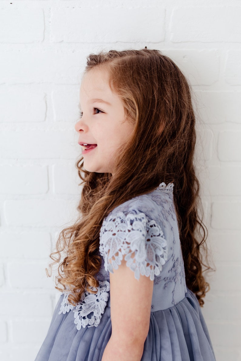 K&ouml;nnte beinhalten: Ein junges M&auml;dchen mit langen braunen Locken tr&auml;gt ein hellblaues T&uuml;llkleid mit Spitzendetails. Das Kleid hat einen gekr&auml;uselten Spitzenbesatz an Ausschnitt und &Auml;rmeln.