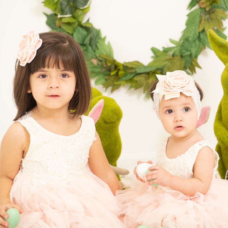 May include: Two young girls wearing pink tulle dresses and white lace tops. The girls are holding pastel colored Easter eggs. One girl has a pink flower headband and the other has a pink flower hair clip.