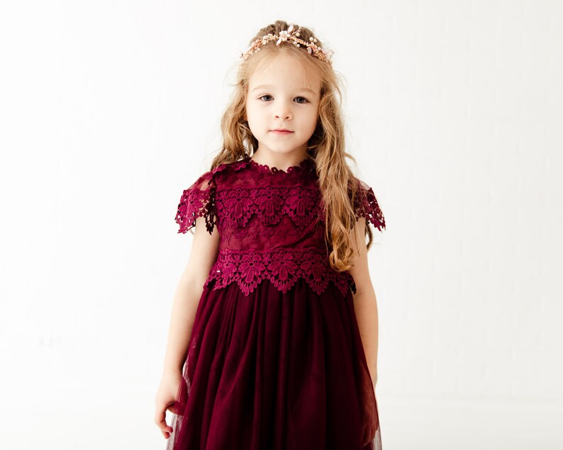 May include: A young girl wearing a burgundy lace dress with a tulle skirt and a floral crown.