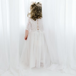 May include: A young girl in a white lace dress with a tulle skirt stands in front of a white curtain, looking out a window.