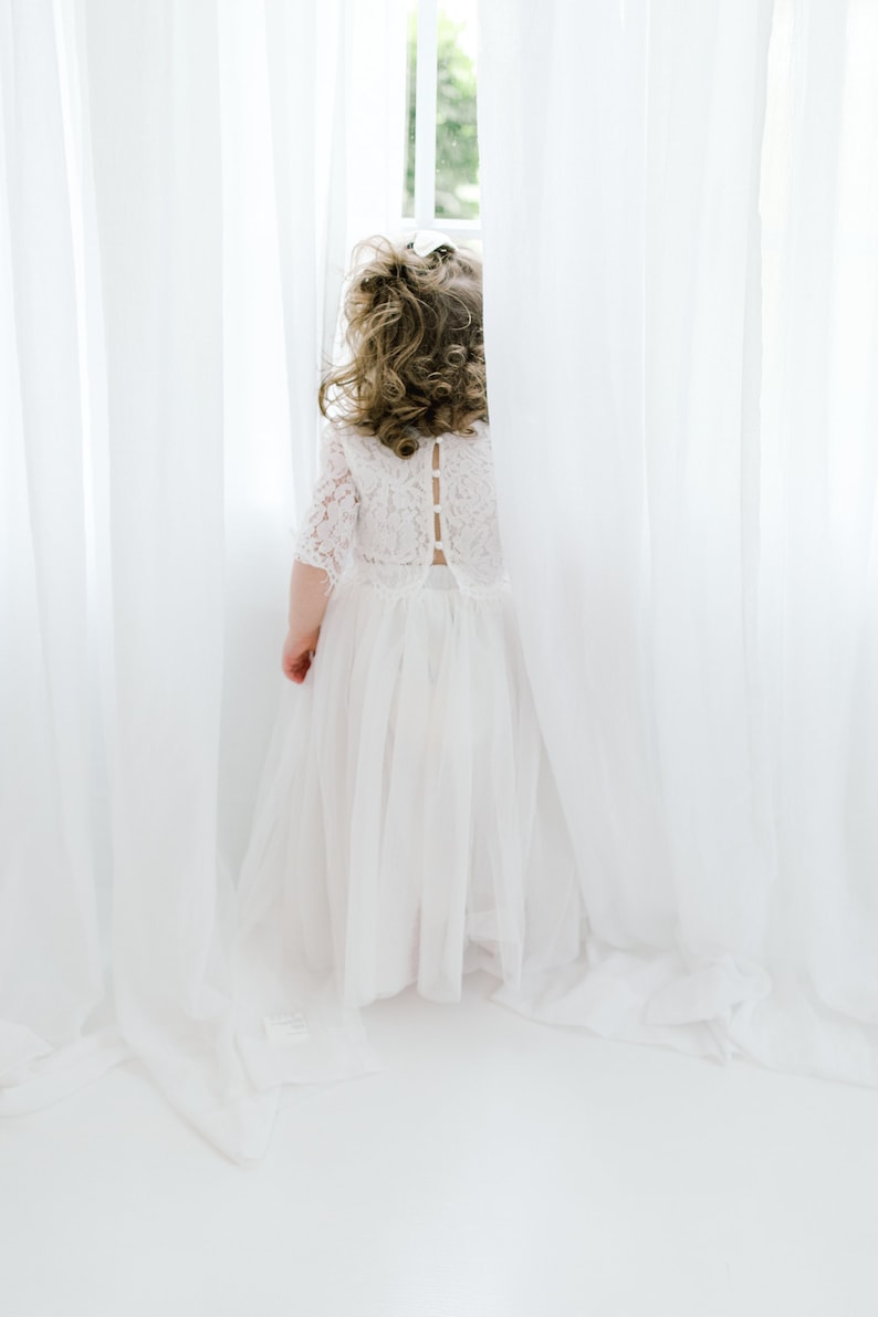 May include: A young girl in a white lace dress with a tulle skirt stands in front of a white curtain, looking out a window.