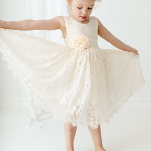 May include: A young girl wearing a white lace dress with a flower accent at the waist. The dress has a scalloped hem and the girl is barefoot.