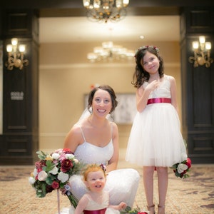 May include: A bride in a white dress with a flower bouquet poses with two flower girls in white dresses. The flower girls are wearing flower crowns and holding bouquets of flowers.