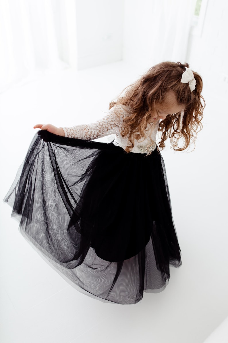 May include: A long-sleeved dress with a white lace bodice and a full, floor-length black tulle skirt. The waist is accented with a decorative embellishment, and the hair is styled with a white bow accessory, creating a formal appearance.