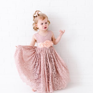 Pu&ograve; includere: Una bambina che indossa un abito di pizzo rosa polvere con una cintura di raso e un fiocco di fiori. L'abito ha una gonna ampia ed &egrave; perfetto per un'occasione speciale.