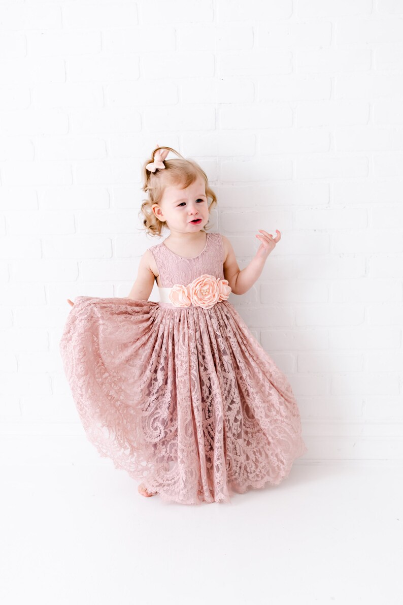 Pu&ograve; includere: Una bambina che indossa un abito di pizzo rosa polvere con una cintura di raso e un fiocco di fiori. L'abito ha una gonna ampia ed &egrave; perfetto per un'occasione speciale.