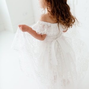 May include: A young girl wearing a white off-the-shoulder lace dress with a satin ribbon tied at the waist. The dress has a flowing skirt and is perfect for a special occasion.