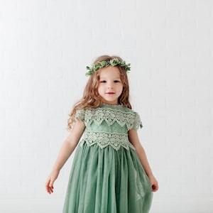 May include: A sage green dress featuring a lace bodice with short sleeves and a scalloped neckline, paired with a flowing tulle skirt. The garment is accented by a delicate floral crown made of green leaves and small white flowers worn on the head.