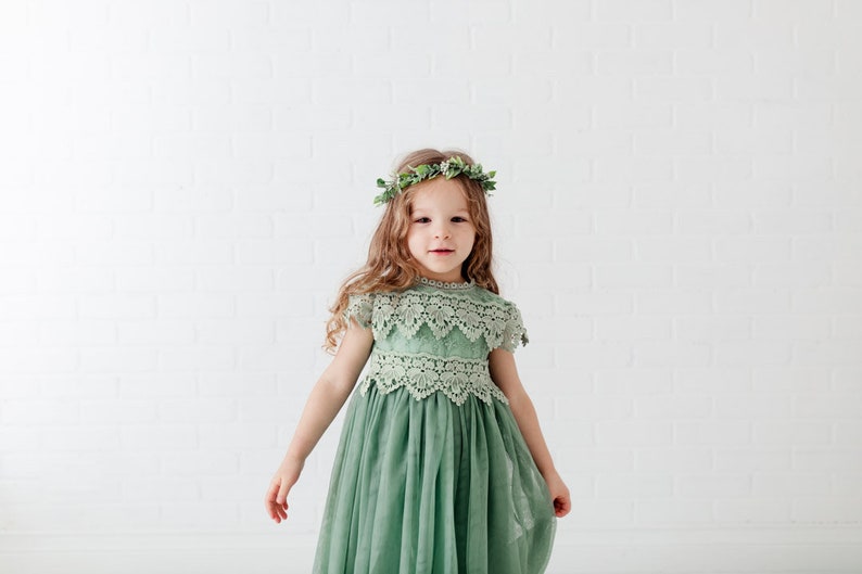 May include: A sage green dress featuring a lace bodice with short sleeves and a scalloped neckline, paired with a flowing tulle skirt. The garment is accented by a delicate floral crown made of green leaves and small white flowers worn on the head.