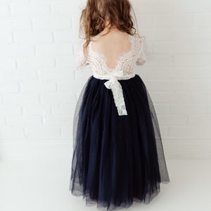 May include: A young girl wearing a white lace dress with a navy blue tulle skirt. The dress has a low back and a white ribbon tied at the waist.