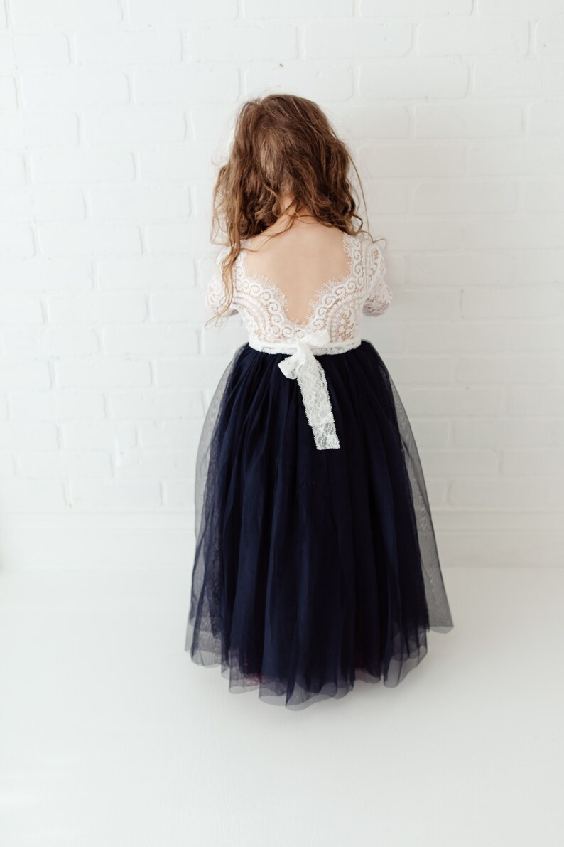 May include: A young girl wearing a white lace dress with a navy blue tulle skirt. The dress has a low back and a white ribbon tied at the waist.