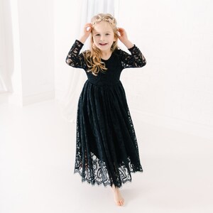 May include: A long-sleeved black lace dress featuring a fitted bodice and a flowing, floor-length skirt with a scalloped hemline. The intricate floral lace pattern covers the entire garment, which has a round neckline and a defined waistline.