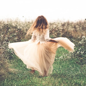 May include: A long dress featuring a white lace bodice with three-quarter length sleeves and a flowing, floor-length beige tulle skirt. The garment is captured in motion, with the skirt flared out to the sides, showcasing the layered fabric.