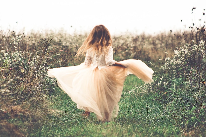 May include: A long dress featuring a white lace bodice with three-quarter length sleeves and a flowing, floor-length beige tulle skirt. The garment is captured in motion, with the skirt flared out to the sides, showcasing the layered fabric.