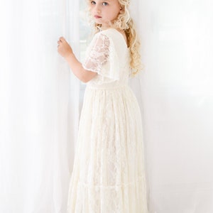 以下が含まれることがあります： 白いレースのドレスを着て、金色の花の冠をかぶった若い女の子。白い背景の前に立っています。