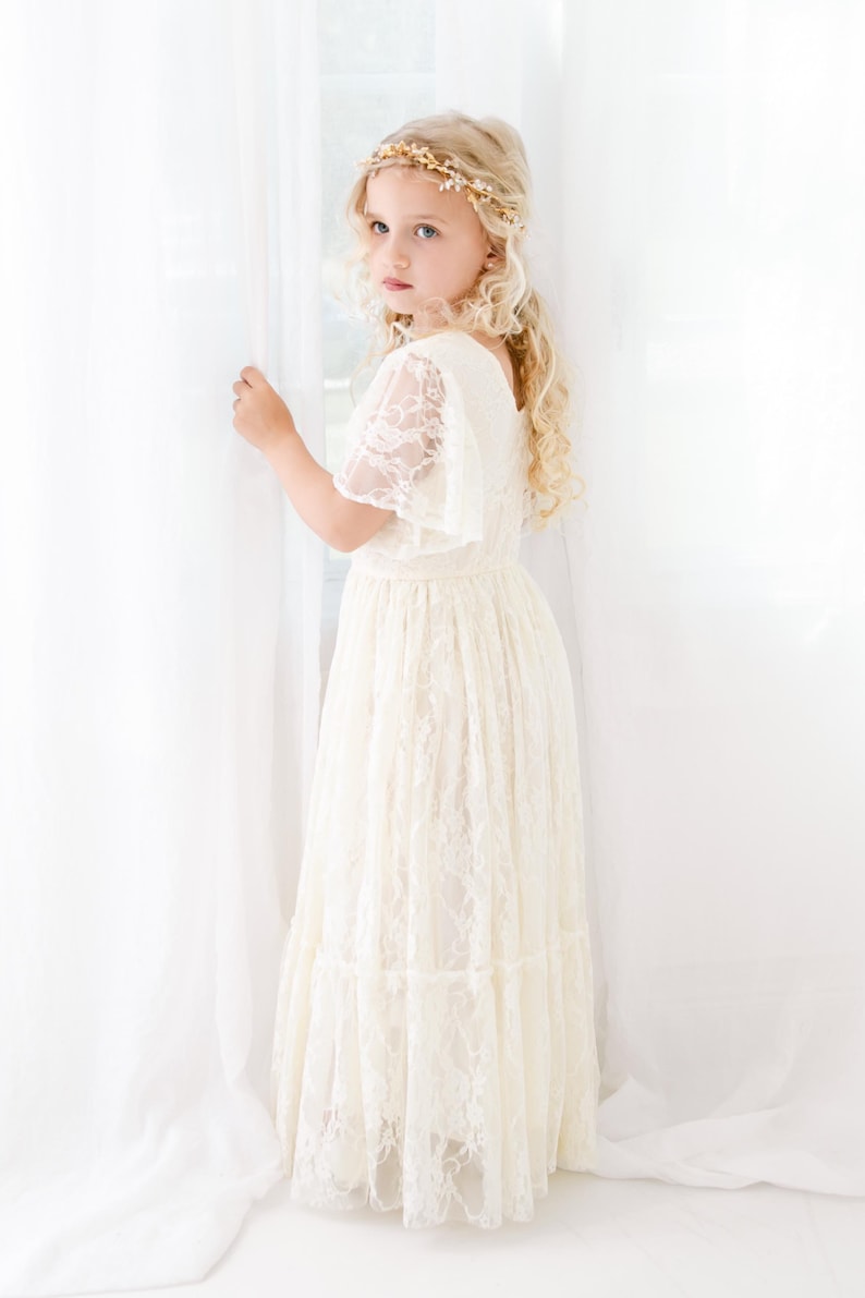 以下が含まれることがあります： 白いレースのドレスを着て、金色の花の冠をかぶった若い女の子。白い背景の前に立っています。