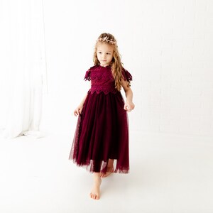 May include: A young girl wearing a burgundy tulle dress with lace detailing and a floral crown. The dress has a fitted bodice and a full skirt. The girl is standing on a white background.