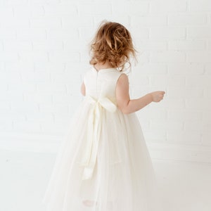 May include: A young girl wearing a white tulle dress with a satin sash. The dress has a sleeveless bodice and a full skirt. The girl is standing in front of a white brick wall.