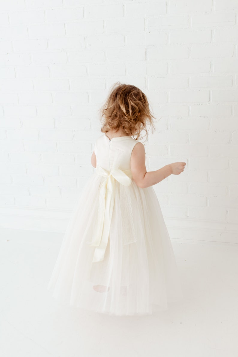 May include: A young girl wearing a white tulle dress with a satin sash. The dress has a sleeveless bodice and a full skirt. The girl is standing in front of a white brick wall.