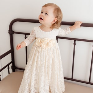 May include: A young girl wearing a white lace dress with a flower sash. The dress has long sleeves and a full skirt. The girl is sitting on a brown metal bench.