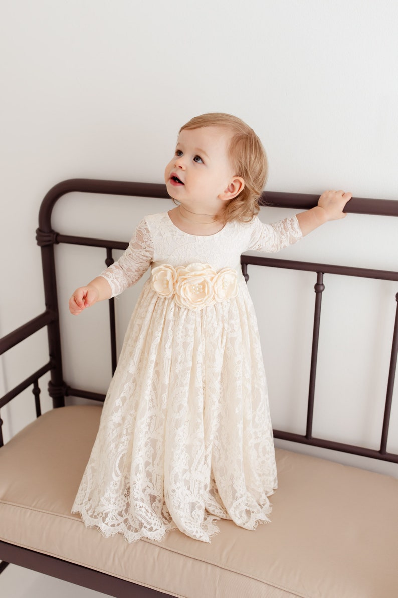 May include: A young girl wearing a white lace dress with a flower sash. The dress has long sleeves and a full skirt. The girl is sitting on a brown metal bench.