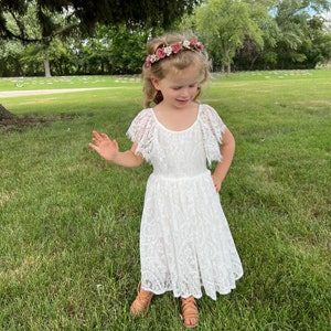 May include: A young girl wearing a white lace dress with a floral crown. She is standing in a grassy area with trees in the background.